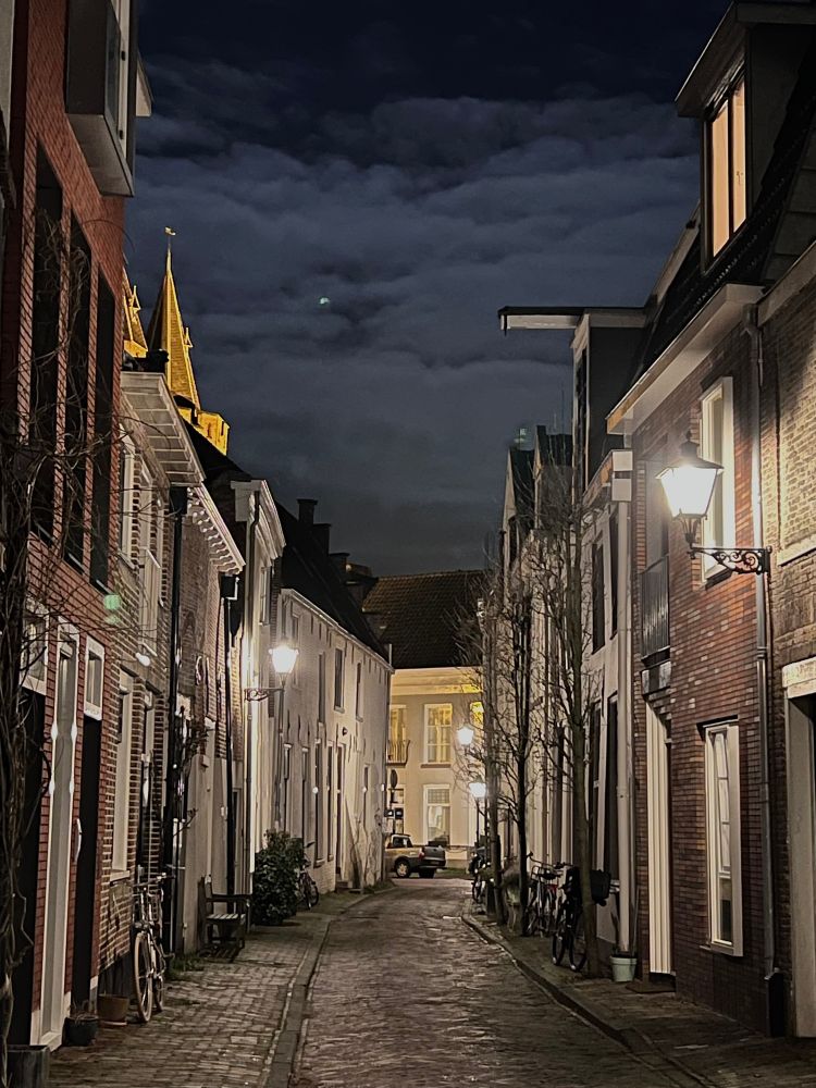 Sfeervolle middeleeuwse straat in de avond, verlicht door klassieke lantaarnpalen. De straat is smal, met ouderwetse bakstenen en witte gevels aan weerszijden. Fietsen staan tegen de muren geparkeerd en er is een parkbankje zichtbaar. Boven de woningen steekt een verlichte kerk- of torenspits uit. De lucht is donker met wolken, wat zorgt voor een gezellige, rustige sfeer. Aan het eind van de straat staat een auto geparkeerd voor een groot, verlicht pand.
