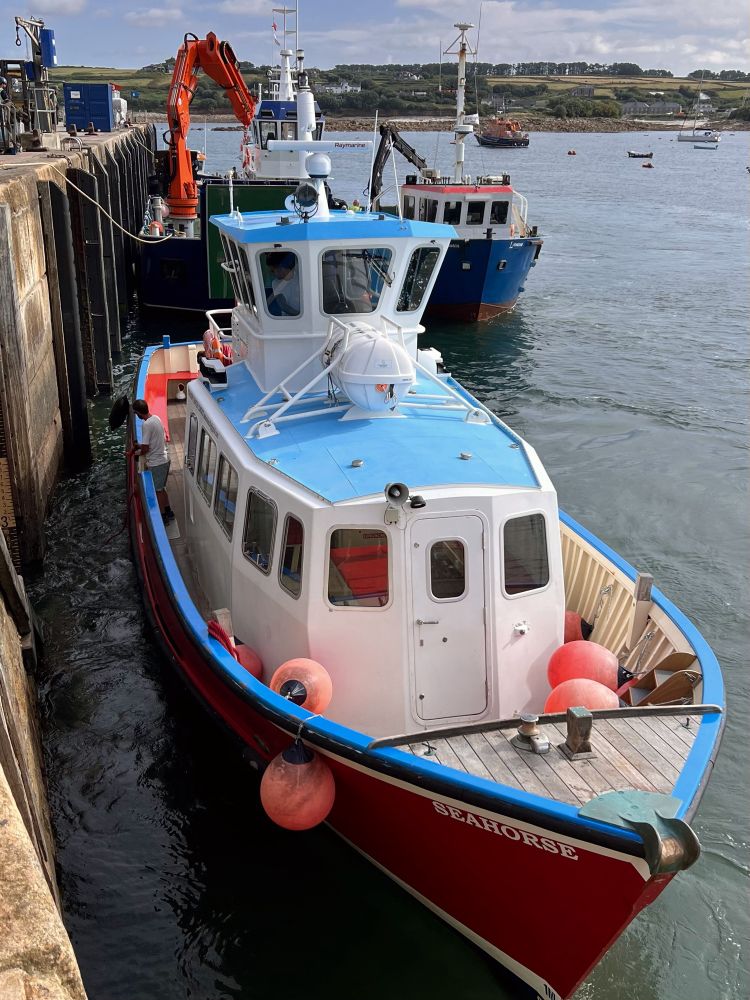 Rode en witte boot met de naam "SEAHORSE" aangemeerd aan een kade naast andere boten. De boot heeft felle blauwe accenten, een houten dek en is voorzien van meerdere oranje drijfballen aan de zijkant als stootwillen. Er staan mensen op de boten en op de kade. Op de achtergrond zie je andere vissersboten en een rustige zee met enkele kleine bootjes en een groene kustlijn. De lucht is licht bewolkt.  