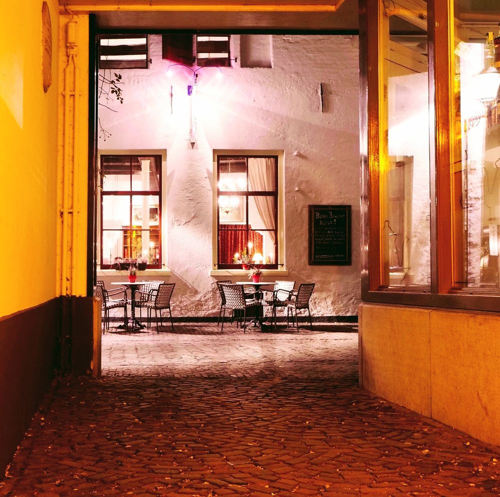 Sfeervol verlichte steeg met zicht op een terras.  
De foto toont een smalle doorgang met kasseien die uitkomt op een charmant terras van een restaurant of café. Aan het einde van de doorgang staan metalen tafels en stoelen, elk tafeltje is gedekt en voorzien van bloemen. De gevel van het gebouw is wit gepleisterd, met twee grote ramen waar warm licht naar buiten straalt. Aan de muur hangt een krijtbord, waarschijnlijk met het menu of een welkomstboodschap. De verlichting, een mix van binnen- en buitenlampen, zorgt voor een gezellige en uitnodigende sfeer. Het is avond of nacht, want er is geen daglicht zichtbaar.