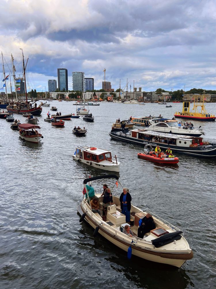 Drukke grachten met veel boten op het water. Op de voorgrond vaart een lichte sloep met zes mensen, onder wie een persoon met een hond. In de achtergrond zijn verschillende soorten boten te zien: kleine motorbootjes, zeilboten, een rode reddingsboot en een grote passagiersboot met veel mensen aan boord. Op de wal zijn moderne hoge gebouwen en enkele kraaninstallaties zichtbaar. De lucht is bewolkt met wat blauwe plekken, het water weerspiegelt de grijze lucht. Er hangt een gezellige en drukke sfeer, waarschijnlijk is er een evenement aan de gang.
