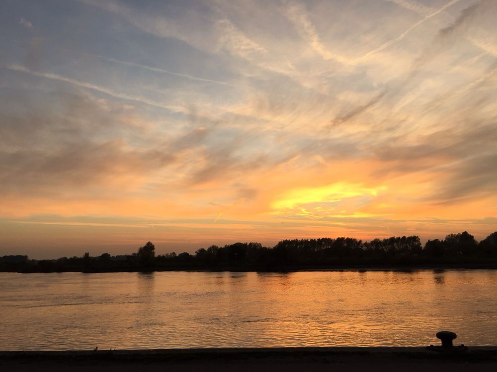 Zonsondergang boven een rivier. Aan de horizon staan donkere silhouetten van bomen. De lucht is gevuld met warme oranje, gele en roze tinten, en er zijn veel dunne, lichte wolken en strepen die diagonaal door de lucht lopen. Het water van de rivier weerspiegelt het gekleurde licht van de zonsondergang. Op de voorgrond is een stukje kade zichtbaar, met rechts een ronde, zwarte aanlegpaal. Het geheel straalt rust uit.
