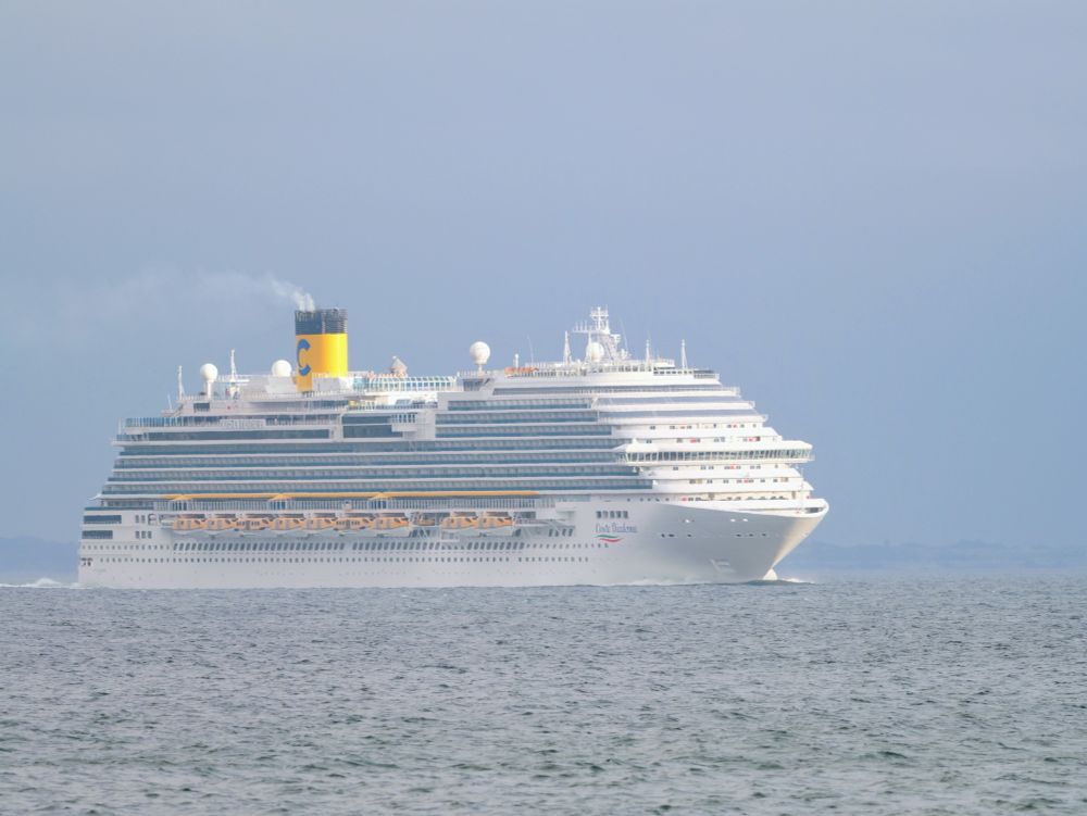 Cruiseschip op zee. Het is een groot wit schip met meerdere dekken en veel ramen, duidelijk gebouwd voor passagiers. Bovenop het schip zijn verschillende satellietschotels en antennes te zien, evenals een grote gele schoorsteen met een blauw logo erop. Aan de zijkant van het schip hangen gele reddingsboten. De naam "Costa Diadema" staat op de zijkant van de boeg geschreven. Het schip vaart op een rustige zee, en op de achtergrond is een licht bewolkte hemel te zien.