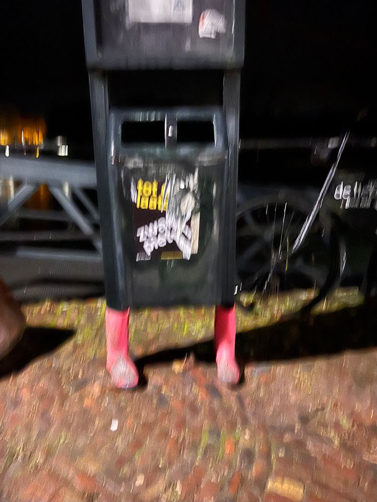 Vuilnisbak, gefotografeerd op een nachtelijke, enigszins onscherpe straat. De vuilnisbak staat op een stenen bestrating en lijkt te worden gedragen door twee objecten die eruitzien als felroze sokken of laarzen, waardoor het lijkt alsof de bak benen heeft. Er zijn fietsen op de achtergrond te zien, evenals een brugleuning. Op de vuilnisbak zit een sticker met de deels leesbare tekst: "tot laat". De sfeer is donker, met wat lichtreflecties in de achtergrond.