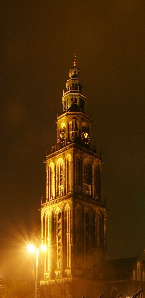 Toren van een kerk, 's nachts verlicht met warme, gele lampen. De structuur is gotisch met hoge ramen en spitsen. Bovenop de toren zit een opengewerkte koepel met een spits. Op de toren zitten twee verlichte ronde klokken. Voor de toren staan twee felle lantarenpalen die licht uitstralen. De hemel is donkerbruin, waarschijnlijk door bewolking of lichtvervuiling.