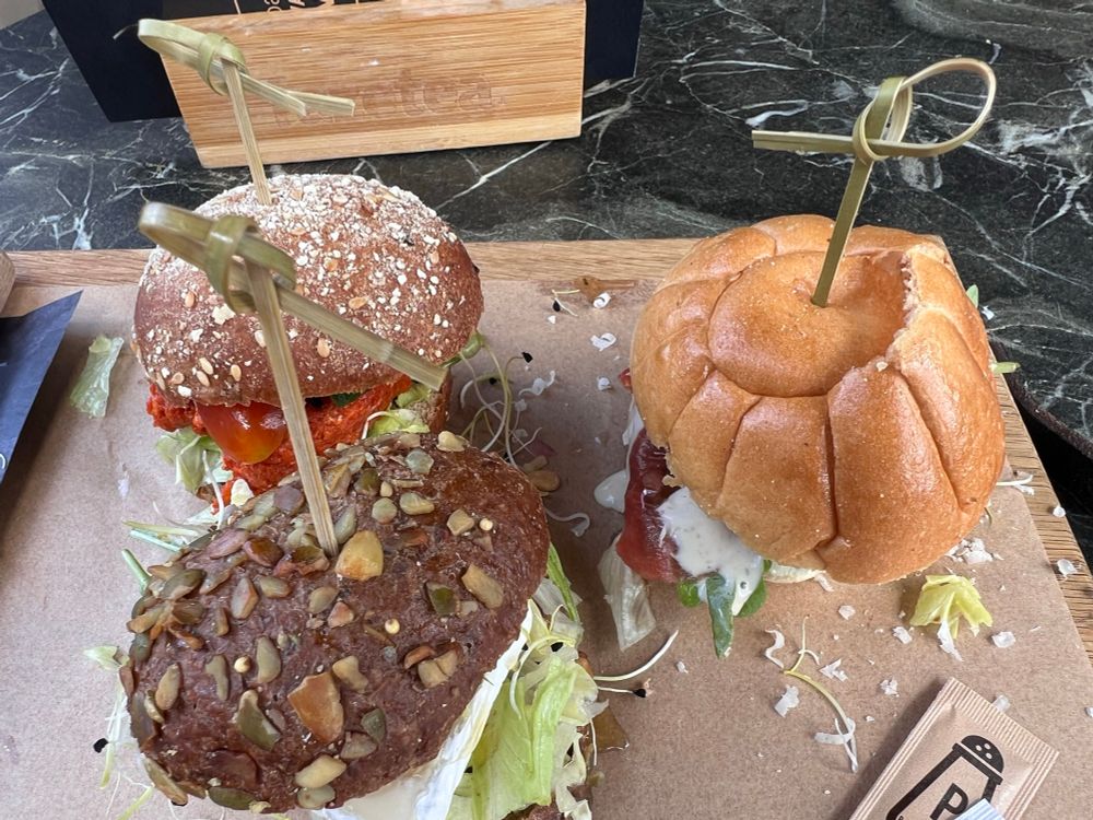 Drie verschillende broodjes op een houten dienblad. Het broodje rechts heeft een zacht, bol wit broodje en bevat sla, tomaat, en een lichte saus. Het broodje linksboven heeft een bruin meergranenbroodje met zaden en lijkt gevuld met een rode, misschien krokante burger (mogelijk vegetarisch of kip), sla en tomaat. Het broodje linksonder is een donkerbroodje met veel pitten en zaden bovenop, gevuld met veel sla en een romige lichte saus. Ieder broodje is vastgestoken met een houten prikkertje en er liggen sliertjes sla verspreid over het dienblad. Op de achtergrond is een zwart met wit marmeren tafel zichtbaar.