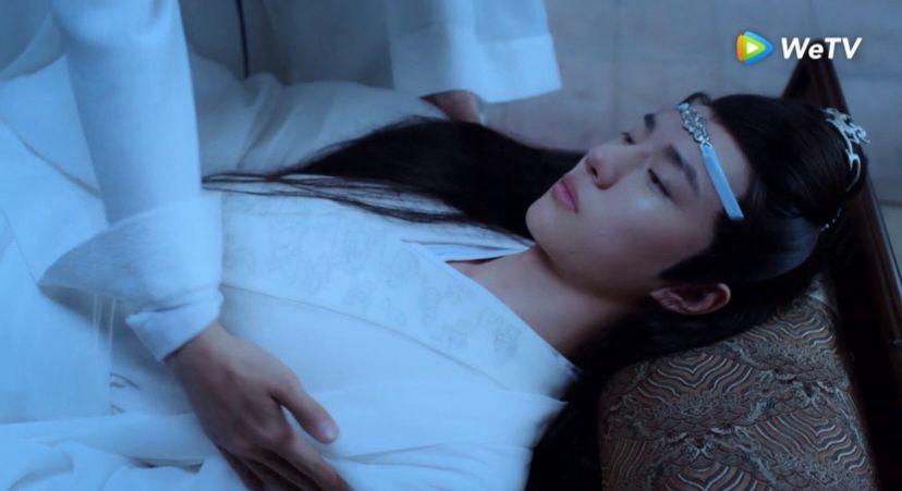 A very drunk teenage Lan Wangji (actor Wang Yibo, dressed in white robes) is guided to lie down on Wei Wuxian's bed with the intent of having Wangji sleep off the alcohol.
The head of LWJ is resting on a patterned pillow while the hands of WWX (actor Xiao Zhan, dressed in white robes) are trying to keep LWJ in place.
