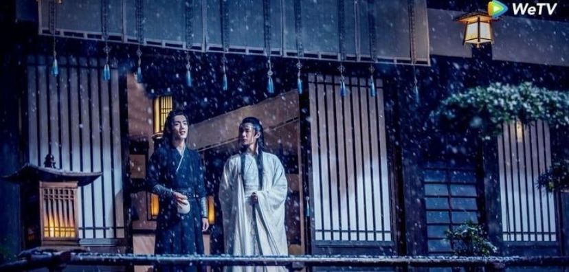 Jingshi while snow is falling.
Wei Wuxian (actor Xiao Zhan, dressed in black robes) and Lan Wangji (actor Wang Yibo, dressed white robes) are standing side by side outside on a wooden deck.
WWX is holding a white jug with ‚Emperor's Smile' in his right hand and is looking straight ahead.
LWJ is holding his left hand in front of his stomach and is looking directly at WWX.

