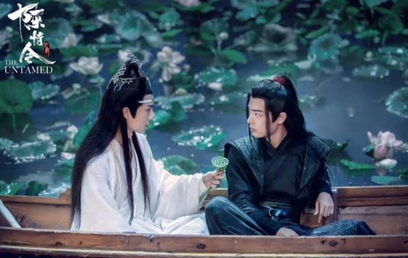 Lotus Lake.
Lan Wangji (actor Wang Yibo, dressed in white robes) and Wei Wuxian (actor Xiao Zhan, dressed in black robes) are sitting next to each other in a wooden boat.
They are looking intensely at each other while LWJ is offering WWX a lotus plant with lotus seeds. 
