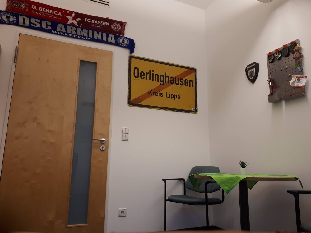 Das Bild zeigt ein Büro  An der Wand hängen zwei Fußballschals von  DSC Arminia Bielefeld und ein Schal vom FC Bayern München.  Ein Straßenschild von Oerlinghausen (Kreis Lippe) ist ebenfalls an der Wand angebracht. Es gibt einen kleinen Tisch mit einer grünen Tischdecke, auf dem sich ein kleiner Topf mit einer Pflanze befindet und zwei Stühle.
