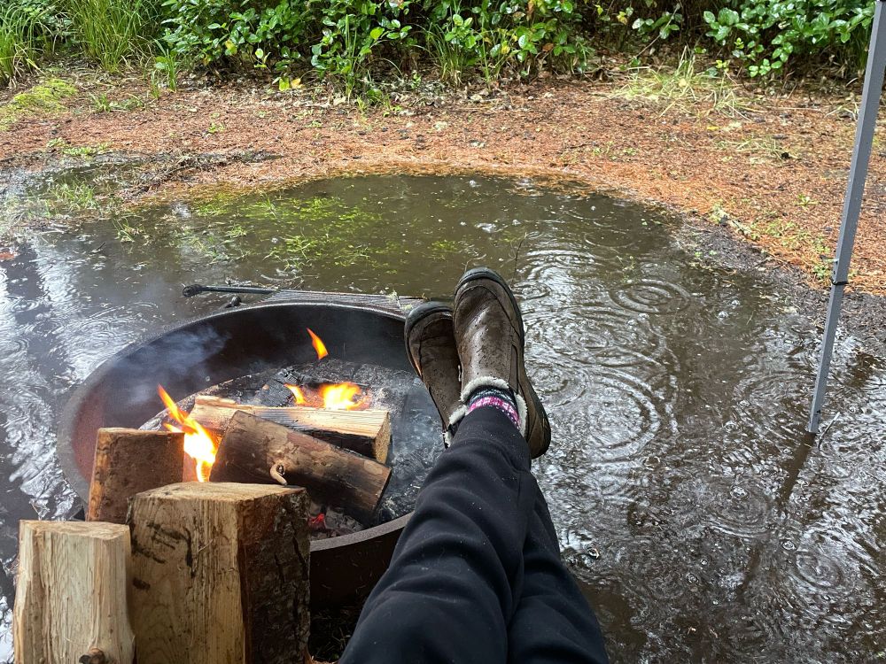 Warming feet near a campfire surrounded by water with wood leaning against the fire pit 