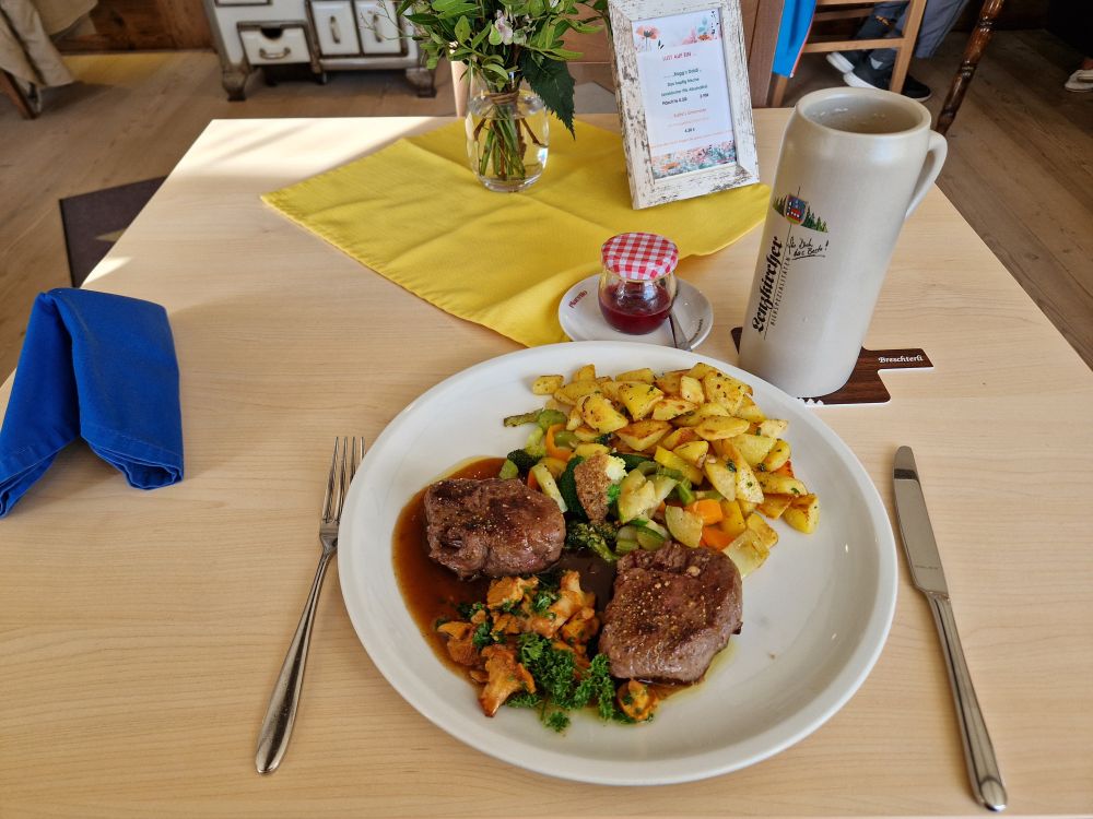 A plate on a table with a yellow cloth on it. On the plate are two venison fillet steaks with mixed vegetables, chanterelle mushrooms and roast potatoes.