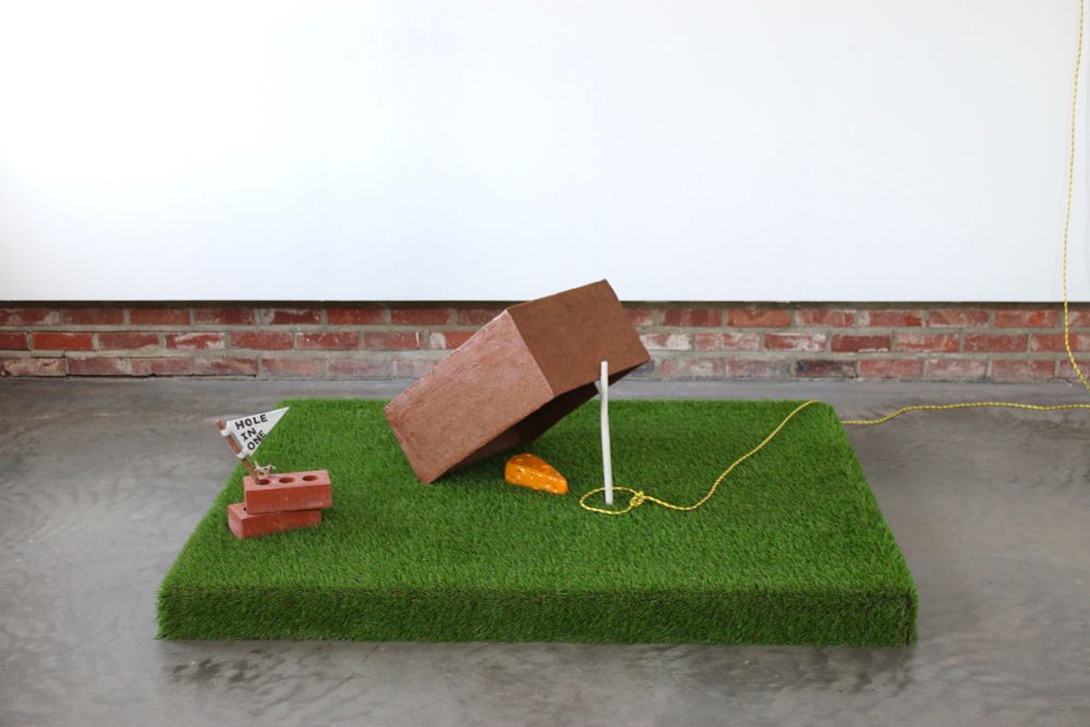 A brown ceramic box lays on top of a rectangle of Astro turf. The box is propped up by a bright white porcelain twig, ceramic cheese is placed under the trap. A yellow rope circles the twig ready to be pulled to catch the prey. 