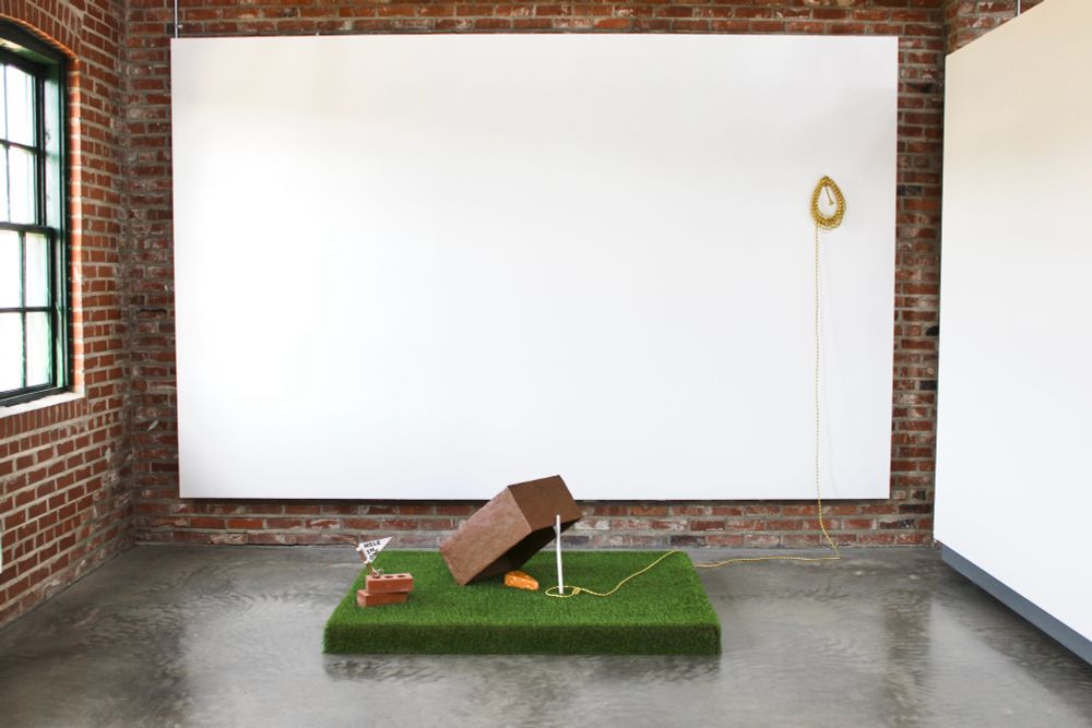 A brown ceramic box lays on top of a rectangle of Astro turf. The box is propped up by a bright white porcelain twig, ceramic cheese is placed under the trap. A yellow rope circles the twig ready to be pulled to catch the prey. 