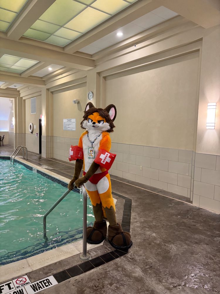 A fox fursuit wearing life guard water wings standing by a hotel indoor pool