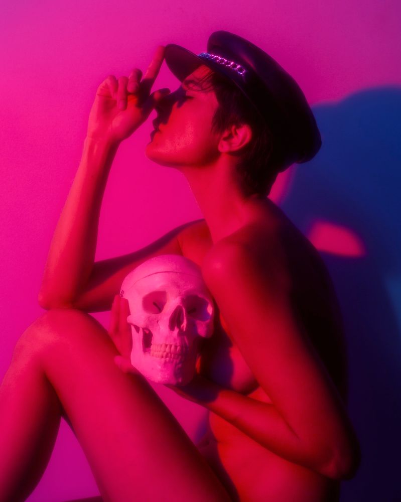 A photo of androgynous model Roaire Yum, wearing a vintage-looking biker cap and holding a skull, while her right hand points to the hat. A blend of red and blue gel light adds pinks and purple to the photo. 