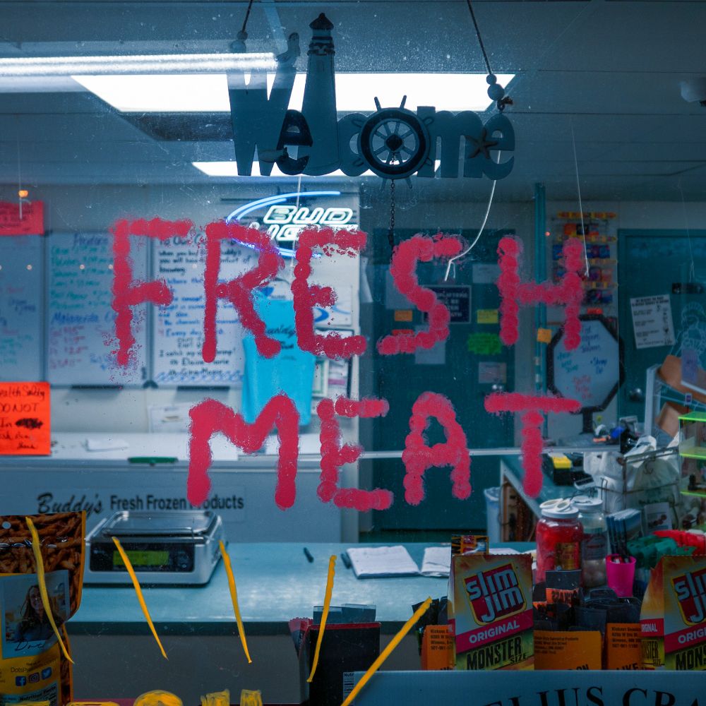 A plate glass window with a "Welcome" sign showing a lighthouse and a wheel for the "o" over the words "FRESH MEAT" daubed onto the window in red paint. 