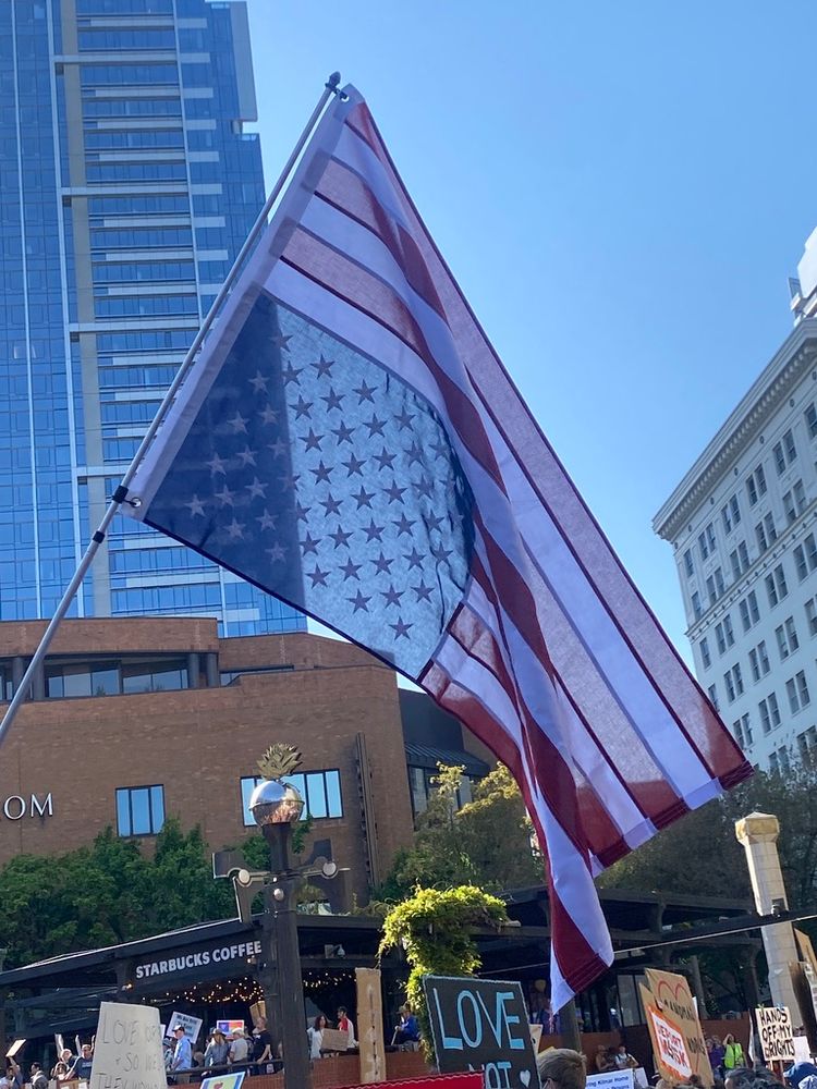 An upside down American flag flies against tall city buildings and a blue sky, above people holding protest signs at the 50501 May Day rally at Pioneer Courthouse Square in Portland, Oregon. 