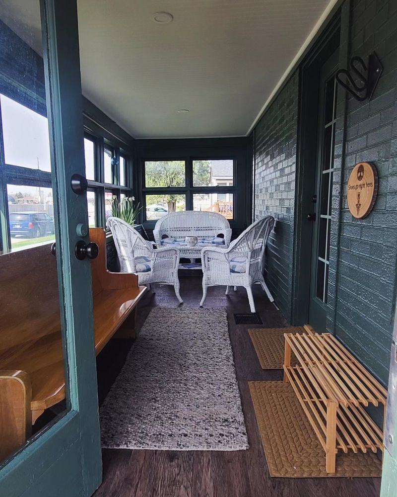 An enclosed porch with windows on the left and green painted faux brick on the right. There's a church pew facing the interior door and a set of white wicker furniture at the end of the space. 
