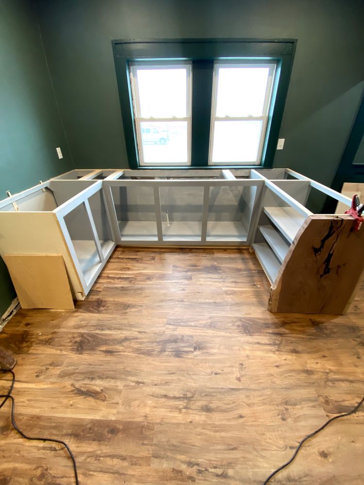 Incomplete kitchen cabinets in front of a window with dark green trim and walls that are a lighter green.