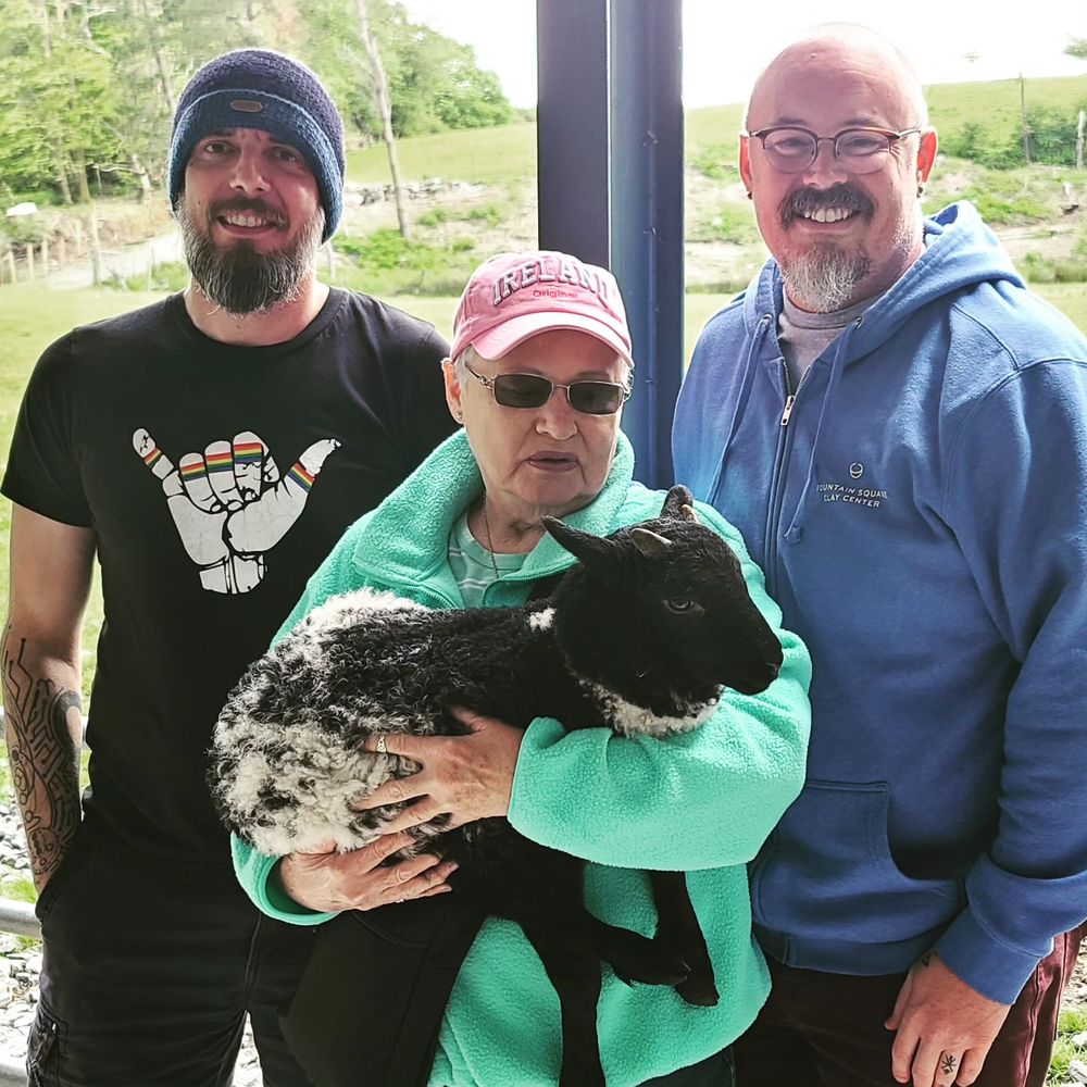Two white men in their late forties stand behind an older white woman who's holding a black and white lamb. 