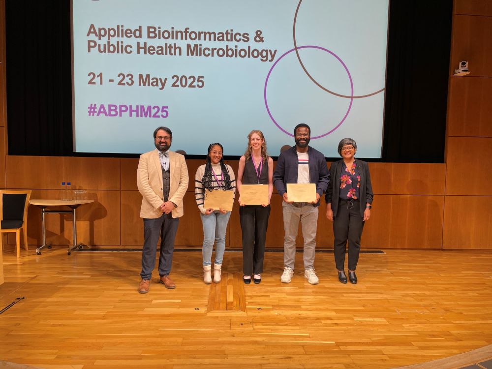 Our two poster prize winners, and one short talk winner for Applied Bioinformatics and Public Health Microbiology accepting their certificates. They are standing on stage within an auditorium, next to two Committee members. They are standing in front of a digital slide carrying the name of the conference. 

Our winners are standing in between our conference Committee members: (LEFT) Nabil-Fareed Alikhan, University of Oxford, UK, and (RIGHT) Senjuti Saha, Child Health Research Foundation (CHRF), Bangladesh