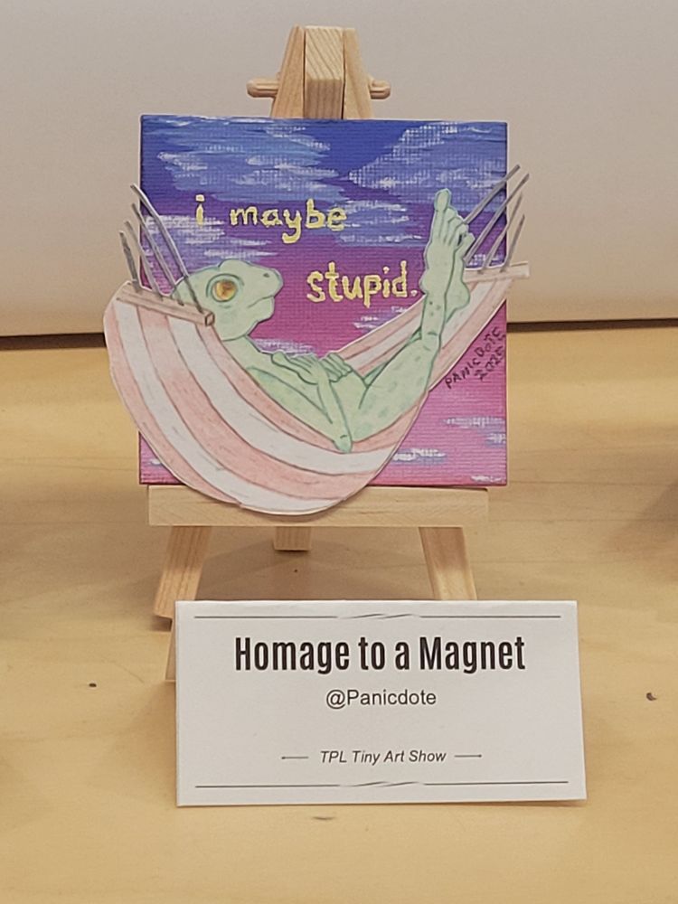 A photograph of a 'Homage to a Magnet' sitting in a little wooden easel. This is a mixed media tiny art piece. A little green spotted frog lies back in a red and white striped hammock, looking up contemplatively with a small frown. The sky above him gathers dusky pinks, purples, and blues, heavy with clouds. The hammock itself escapes a bit from the confines of the 3x3 inch canvas. In pale yellow lettering, text appears around the frog's head. In all lowercase, the text reads, "i maybe stupid." The piece is signed in small black text: Panicdote 2025.
