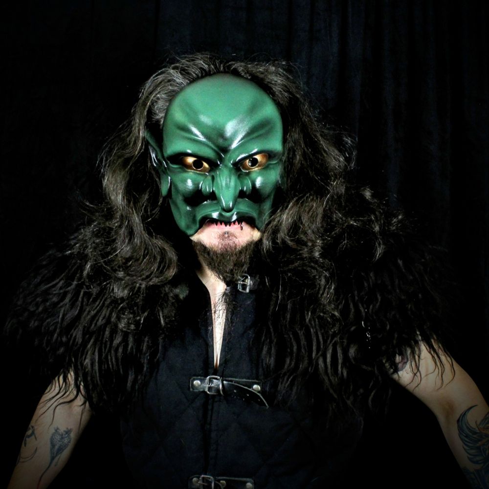 A model wearing a goblin mask with long dark hair and a fur mantle glares directly at the viewer