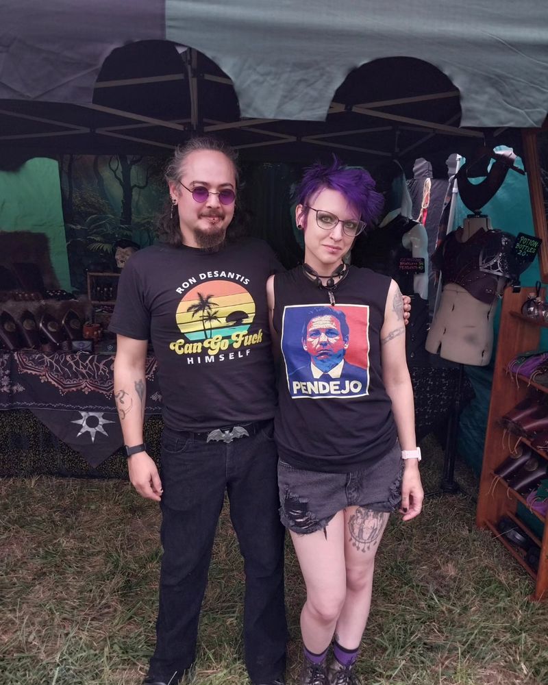 A man and woman standing outside in front of a vendor booth. Both are wearing anti-ron desantis shirts.