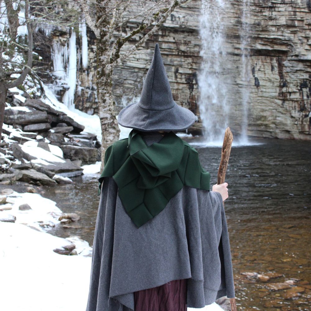 A model with their back to the viewer in layers of wool cloaks and a grey pointy wizard hat. The model holds a driftwood staff and faces a waterfall