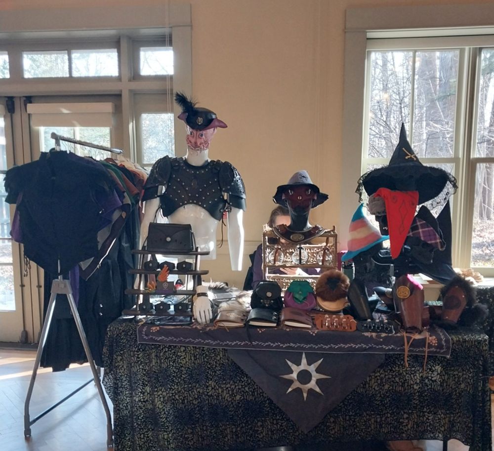 A table with green and black tablecloths full of leather goods, a hat rack and a clothing rack on the left