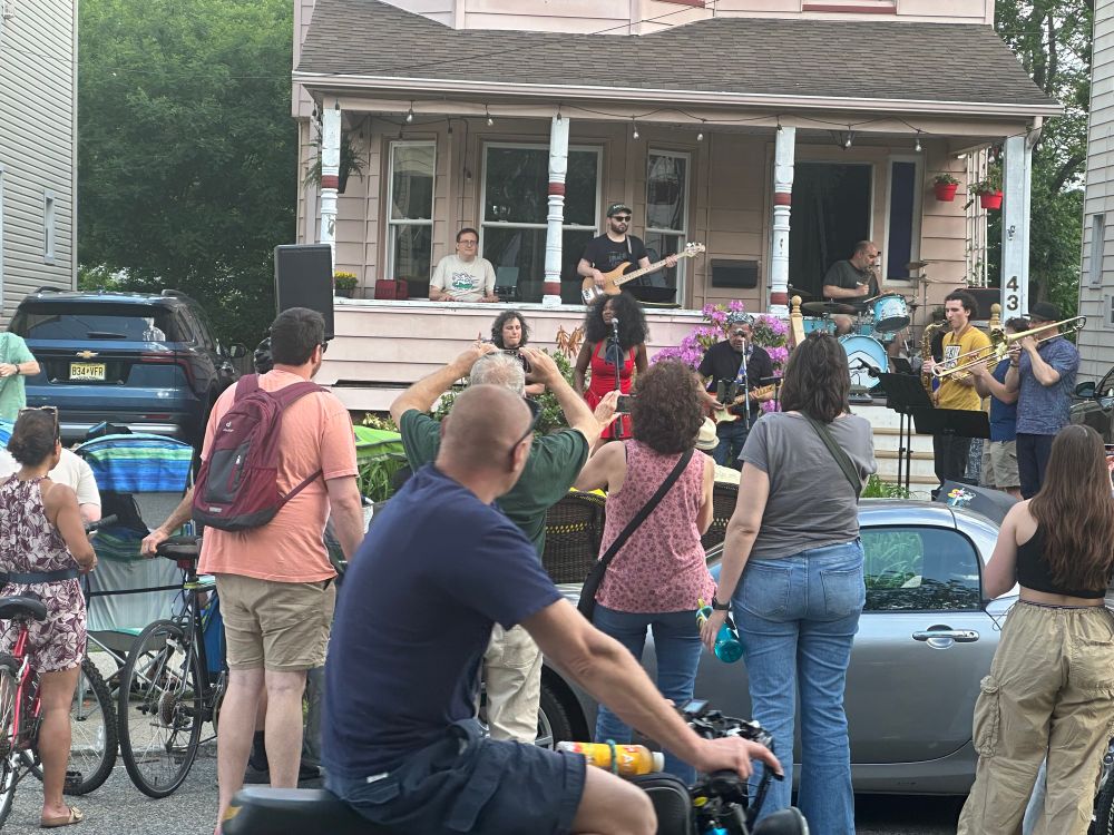 A band plays on the lawn of a suburban house. The keyboard, bass, & drummer are on the porch. The two singers and guitar are on the lawn in front of the porch. A 3-piece horn section is off to the right.
People are standing next to parked cars or on the lawns. A man on a bike rolls past looking at the band.