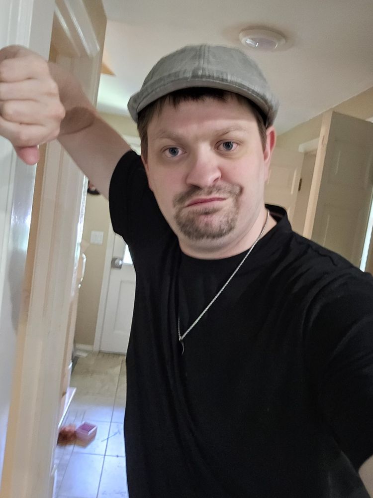 Me looking hot af if I do say so myself - which, I do - in a black t-shirt, silver necklace, and a grey flat cap. I'm leaning against a door and honestly need to shave. 