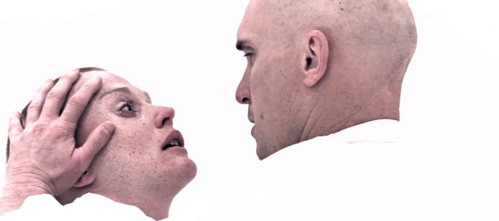 A male and a female, bald, wearing white coveralls, in an all-white environment, with distraught expressions, embracing. A still from Geo. Lucas' first feature film, THX 1138.