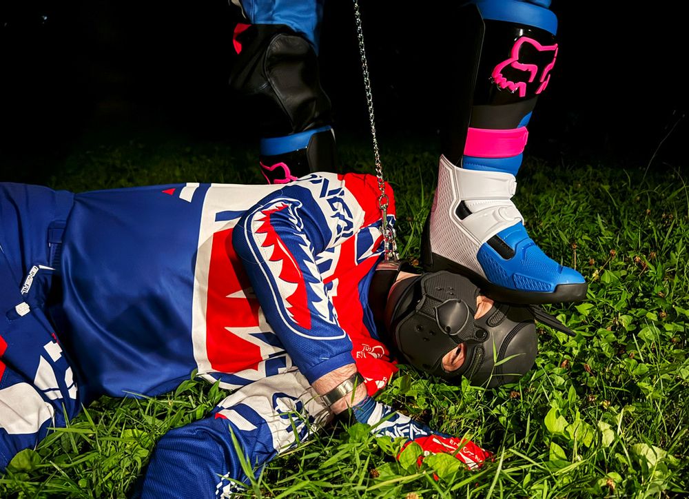 max grabbing me by the leash and stepping on my head. we're both wearing red white and blue motocross gear. I'm in a black pup hood