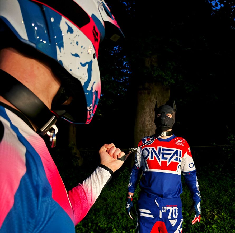 max grabbing me by the leash. we're both wearing red white and blue motocross gear. I'm in a black pup hood