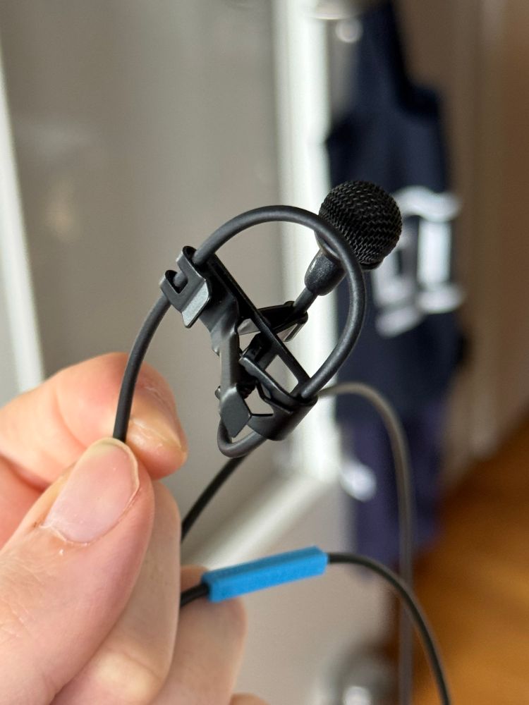 Nahaufnahme eines Sennheiser MKE-2 Ansteckmikrofons. Zu sehen ist wie das Kabel in einer Art Schlaufe innerhalb der mitgelieferten Halteklammer geführt wird.