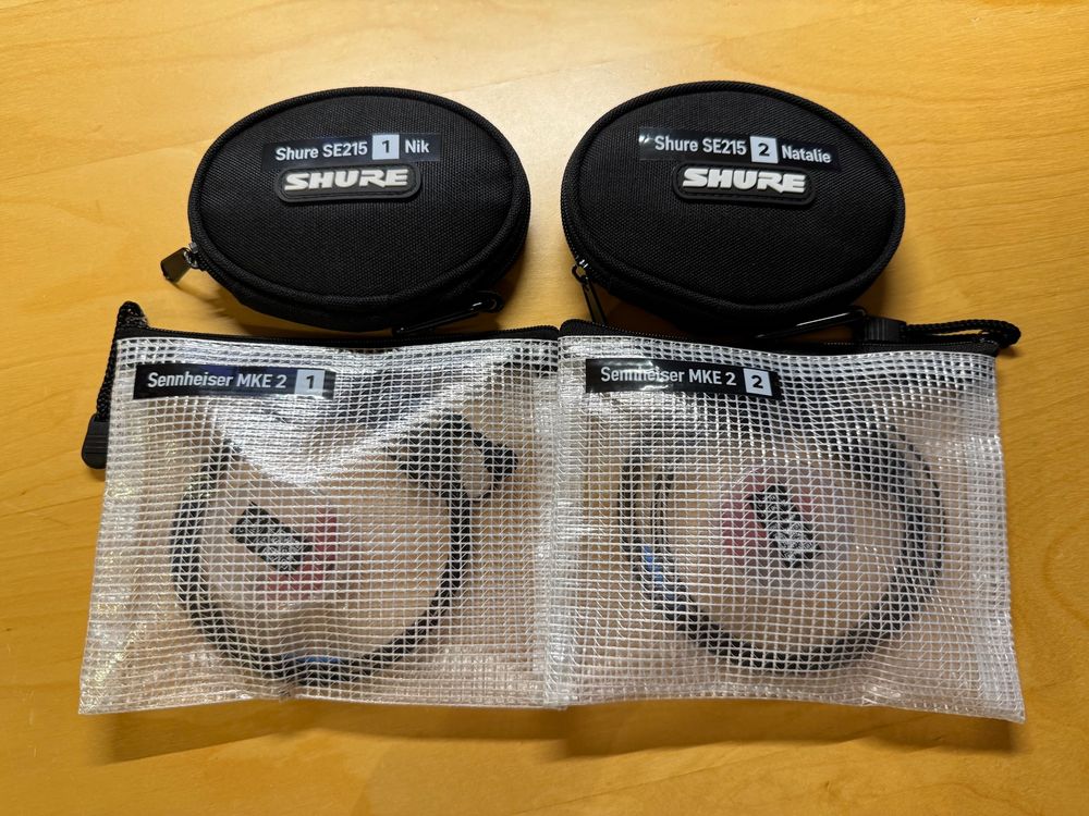 Foto von zwei Transporttaschen-Paaren. Im oberen befinden sich Shure SE215 In-Ear-Monitorkopfhörer, im unteren sind Sennheiser MKE 2 Lavaliermikrofone verstaut. Alle Taschen sind entsprechend mit ihrem Modellnamen und einer Ziffer zur Differenzierung auf einem schwarzen Label mit weißem Text beschriftet. Das Taschenpaar mit den Ohrhörern ist zusätzlich mit den Namen „Nik“ und „Natalie“ neben Modellbezeichnung und Nummerierung beschriftet.