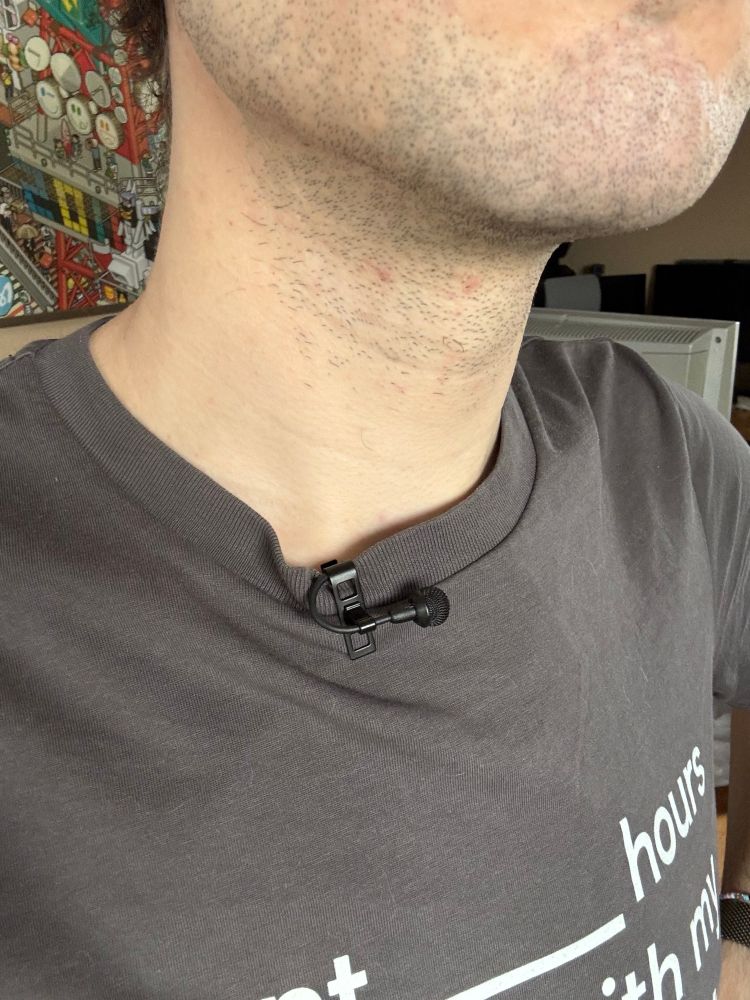 Nahaufnahme eines Sennheiser MKE-2 Ansteckmikrofons am Kragen eines T-Shirts. Das Kabel wird hier ebenso innerhalb der Klammer hinter das Shirt weggeführt.