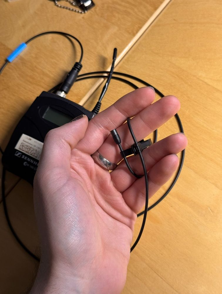 Eine Hand hält ein kleines schwarzes Lavaliermikrofon mit Clip, das an ein Kabel angeschlossen ist. Im Hintergrund liegt ein drahtloser Sennheiser-Sender auf einem Holztisch.