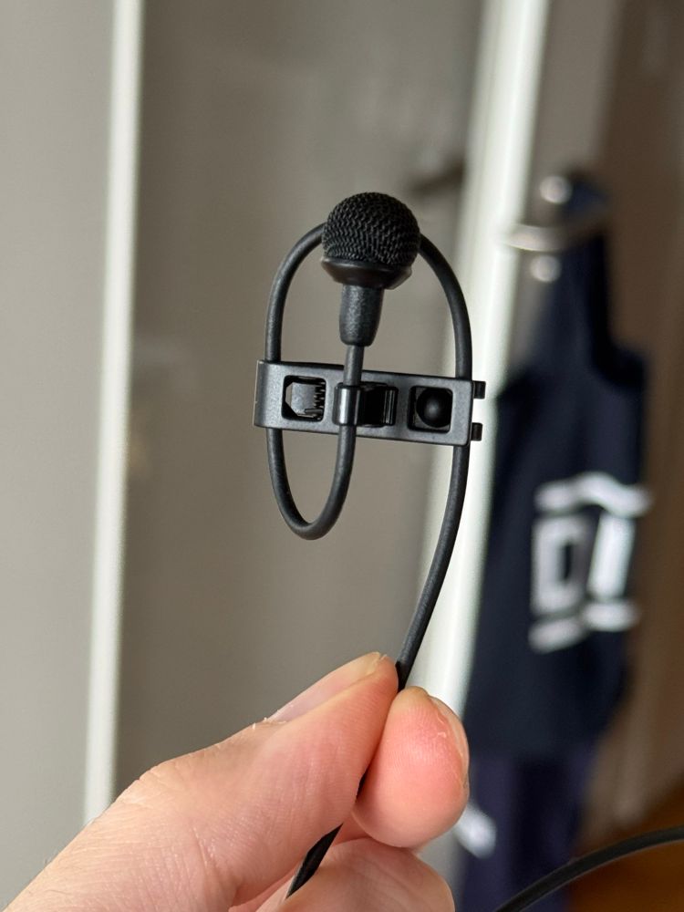 Nahaufnahme eines Sennheiser MKE-2 Ansteckmikrofons. Zu sehen ist wie das Kabel in einer Art Schlaufe innerhalb der mitgelieferten Halteklammer geführt wird.
