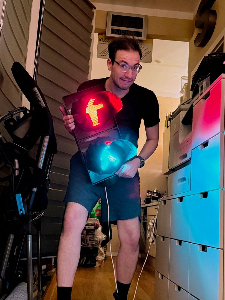 Autor in schwarzem T-Shirt und Shorts hält eine alte Fußgängerampel mit leuchtendem roten DDR-Ampelmännchen oben und grünem unten, beide gleichzeitig eingeschaltet.