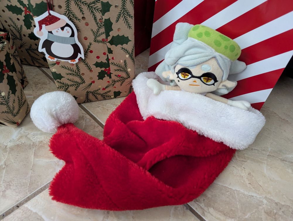Marie Plush is wrapped inside a Santa hat, with only her head and arms poking out. She lays on the floor, looking at the camera, surrounded by a few presents.
