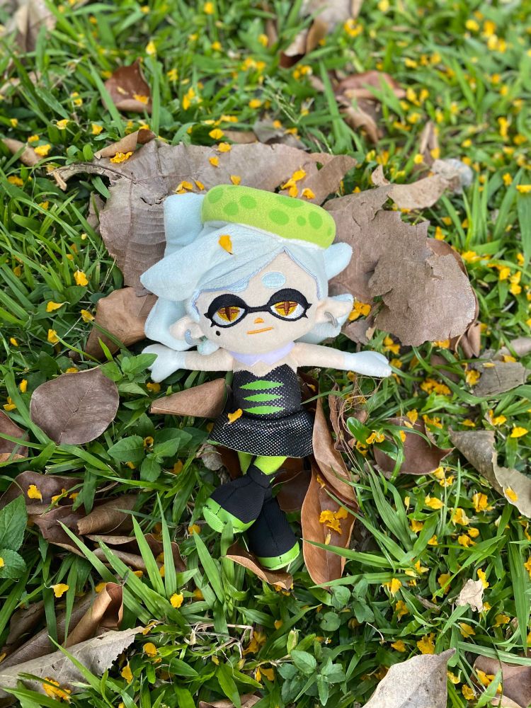 Marie Plush is relaxing on the grass, arms wide open and legs crossed, surrounded by dry leaves and small yellow flower petals. The photo was taken from a top-down perspective with Marie on center focus on a partly cloudy afternoon.