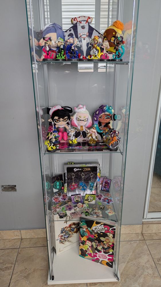 A glass cabinet with three selves filled with Splatoon merchandise (plus a couple of extras). 
The top shelf has the three Deep Cut plushies and the seven idol amiibos. 
The shelf below has Callie Plush, the two Off the Hook plushies, six amiibo (four alternate idols, S1 inkling boy, Link (TotK)), and the S3 Grand Festival wristband.
The shelf below is my lil Marie shrine, and thus features a lot of Marie (with a hint of Callie): eight keychains, three enamel pins, and four stickers. This shelf also has a folded S3 bag and TotK keychain, and the Splatoon x Zelda splatfest keychains from My Nintendo, and some extra cards and stickers with Splatoon characters. 
The floor of the cabinet houses two artbooks: Zelda BotW Creating a Champion, and The Art of Splatoon 2.