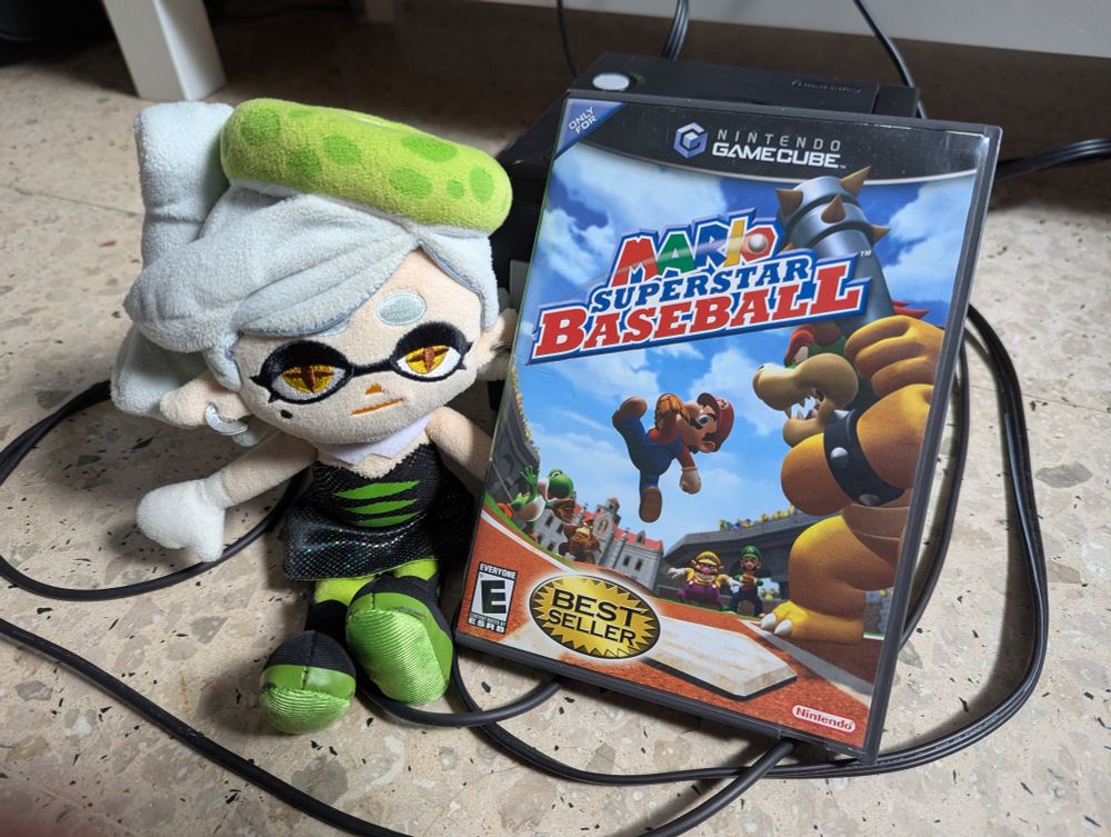 A physical copy of Mario Superstar Baseball is propped in front of a GameCube console and beside Marie Plush.  