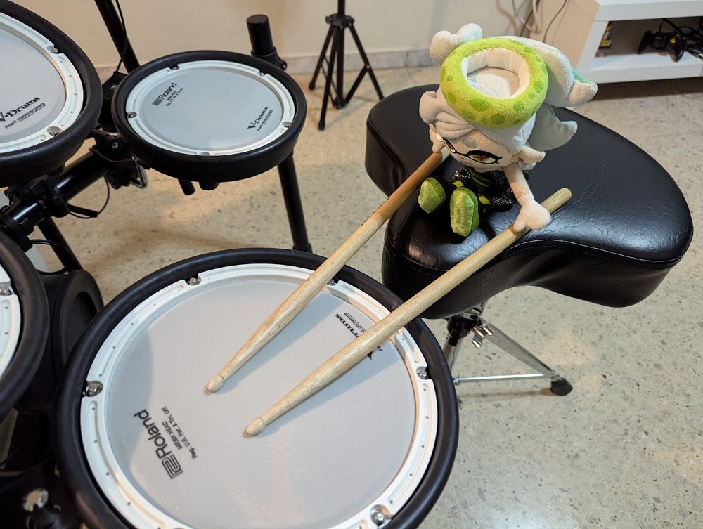 Marie Plush plays on an electronic drum kit. She sits on a drum stool positioned in front of the snare drum, with drumsticks placed by her hands with the tips/shoulders of the sticks resting on the drum pad (as if she's holding them). The photo was taken from a top-down, 3/4 perspective, looking from the left side of the kit toward the right side. 