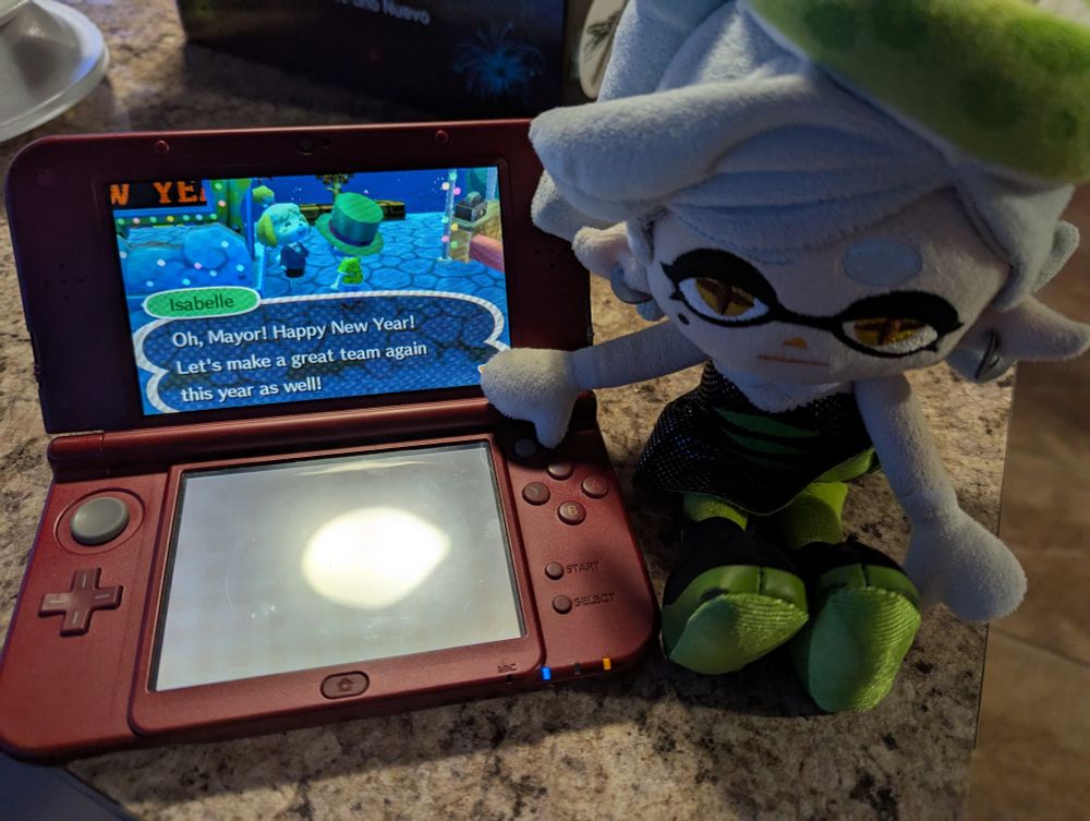 Marie Plush posing next to a New 3DS playing Animal Crossing New Leaf. In-game, my player character is at the plaza celebrating the New Year. Isabelle's dialogue reads: "Oh, Mayor! Happy New Year! Let's make a great team again this year as well!"