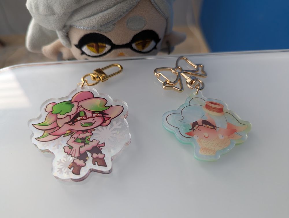 A 2.5-inch acrylic keychain featuring Marie in a holiday outfit, and a 2.5-inch pearlescent charm of FrostyFest Marie. The two items are being displayed on a table; Marie Plush's head peeks over the table to look at the items. 