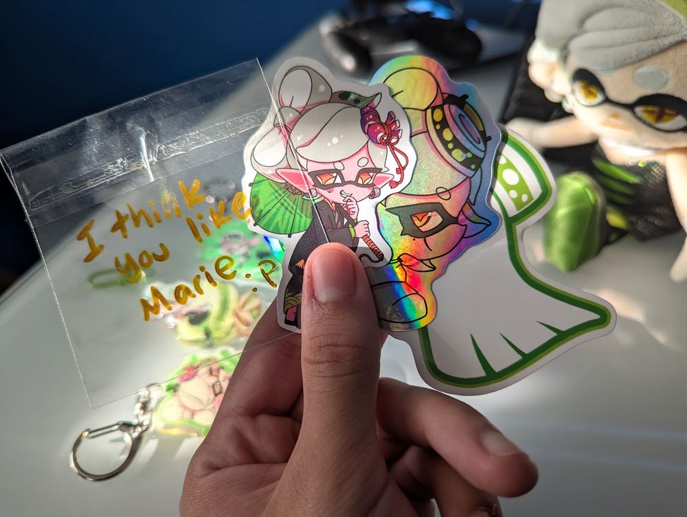 I'm holding in my hand three stickers and the bag they came in. The stickers are: Marie in her kimono outfit, a holographic sticker of Marie in her Alterna outfit giving a side eye to the viewer, and Marie's symbol from the Squid Sisters logo. The plastic bag has text written on it saying "I think you like Marie :P".