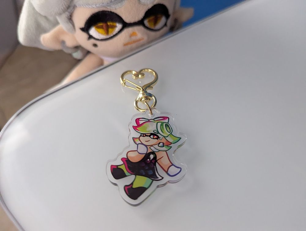 A 2-inch acrylic charm featuring Marie in her standard outfit. Marie Plush is lurking in the background looking at the charm. 